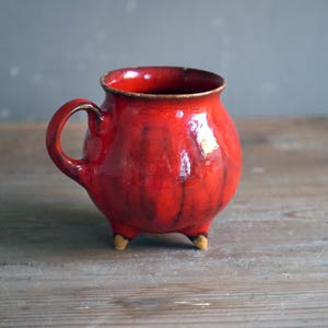 May include: A handmade, red ceramic mug with a glossy finish. The mug has a rounded body, a handle, and three small feet. The interior and rim are a darker shade of red, and the mug sits on a wooden surface.