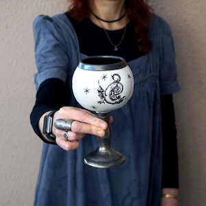 May include: A white ceramic goblet with a black dragon illustration and star accents. The goblet has a silver-colored stem and rim. The dragon design is detailed and stylized.