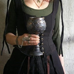 May include: A silver chalice with a detailed, textured design is held in front of a person wearing a black corset-style top over a green long-sleeved shirt. The chalice has a long stem and a wide bowl. The person is also wearing rings and a bracelet.
