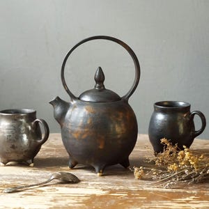 May include: A rustic tea set featuring a dark, metallic-finished teapot with a large handle and two matching mugs. The teapot and mugs sit on a wooden table with a spoon and dried flowers. The set has a handmade, artisanal aesthetic.
