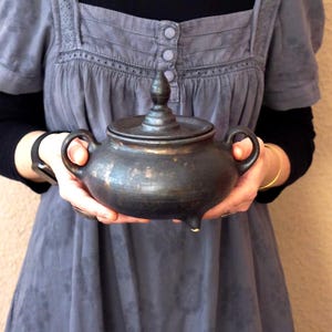 May include: A dark, handcrafted ceramic pot with a lid, two handles, and three small feet. The pot has a matte black finish with subtle bronze highlights. The lid is topped with a decorative finial. A unique piece for kitchen or home decor.