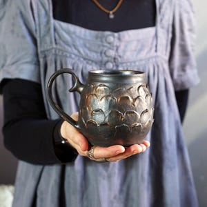 Könnte beinhalten: Eine dunkle Keramiktasse mit einem strukturierten, schuppenartigen Muster. Die Tasse hat einen großen Henkel und eine dunkle, matte Oberfläche. Die Tasse wird in den Händen einer Person gehalten.