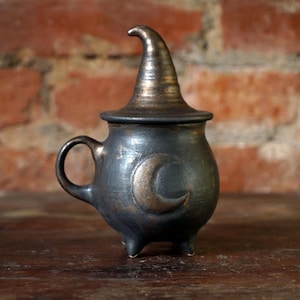 May include: A dark grey ceramic cauldron with a witch hat-shaped lid. The cauldron has a handle, three small feet, and a crescent moon design. The background is a brick wall and a wooden surface.