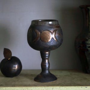 May include: A dark, metallic goblet with a triple moon design, a dark apple with a leaf, and a green vase. The goblet is approximately 6 inches tall. The objects are displayed on a shelf.