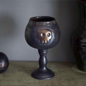 Peut inclure: Un gobelet métallique foncé avec un emblème de crâne, une pomme décorative et un vase en céramique. Le gobelet a un bol arrondi et une fine tige. Le crâne est de couleur or. La pomme a une feuille. Le vase est vert et bleu.