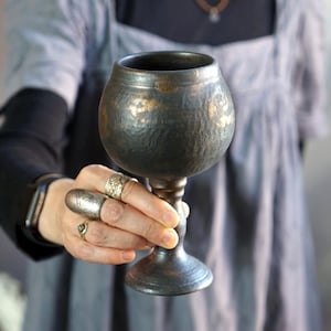 Könnte beinhalten: Ein gehammerter Metallkelch mit dunkler, gealterter Oberfläche und goldenen Akzenten. Der Kelch hat eine abgerundete Schale und einen schlanken Stiel. Der Kelch wird in der Hand einer Person gehalten, mit mehreren Ringen an den Fingern.
