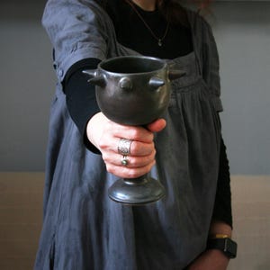 May include: A dark gray ceramic chalice with a flared base and a rounded bowl. The chalice has three conical protrusions around the rim. The stem is smooth and the overall design is rustic and handmade.