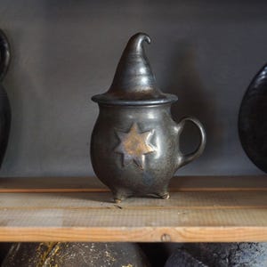May include: A dark gray ceramic vessel with a handle, lid, and three small feet. A gold star is centered on the front. Other ceramic pieces are visible on a wooden shelf.