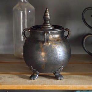 Peut inclure: Un récipient en céramique gris foncé en forme de chaudron avec un couvercle et trois petits pieds. Le récipient a deux poignées sur les côtés et un bouton décoratif sur le couvercle. Le chaudron a une finition dorée métallique.