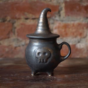May include: A dark gray ceramic cauldron with a witch hat-shaped lid. The cauldron has a skull design on the front, a handle, and three small feet. The background is a brick wall.