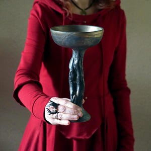 May include: A dark, ornate chalice with a wide, bronze-colored rim and a textured, black stem. The chalice is held against a red hooded garment, creating a striking contrast. The stem has a unique, organic design.