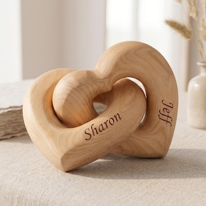 Könnte beinhalten: Zwei ineinander verschlungene Holzherz-Skulpturen mit den eingravierten Namen "Sharon" und "Jeff". Das helle Holz hat eine natürliche Maserung. Die Herzen sind auf einer neutralen Oberfläche platziert.