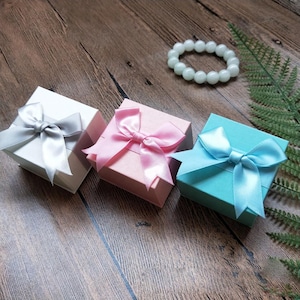 May include: Three small gift boxes in white, pink, and blue, each tied with a matching satin ribbon bow. A light green beaded bracelet and fern frond are also visible on the wooden surface. These boxes are ideal for jewelry or small gifts.