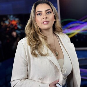 Könnte beinhalten: Eine Frau in einem cremefarbenen Blazer über einem hellen Oberteil hält einen Stapel weißer Karten. Der Hintergrund ist verschwommen mit blauen und bunten Lichtern. Der Blazer hat ein zweireihiges Design.