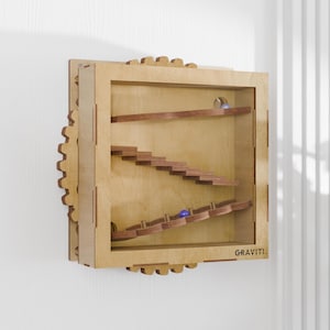 May include: Wooden marble run game with a clear front panel. The game features a series of wooden ramps and stairs for marbles to travel down. The word "GRAVITI" is printed on the bottom right. The game is mounted on a wall.