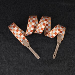 May include: A camera strap with a checkered pattern in orange, white, and blue. The strap has brown leather ends.