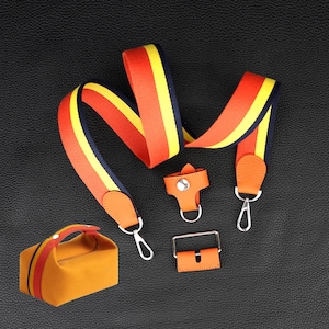 May include: A camera strap with orange, yellow and black striped webbing. The strap has a leather adjustable buckle and a metal hook. The strap is on a black surface.