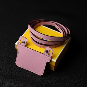 May include: A pink leather strap with a matching pink leather card holder. The strap is wrapped around a yellow box.