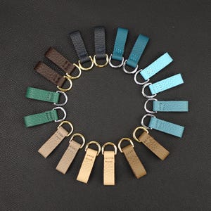 May include: A set of 15 leather key fobs in various shades of blue, green, and brown. Each fob has a silver metal ring.