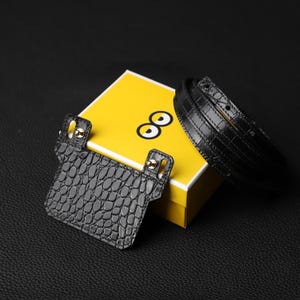 May include: A black crocodile leather belt with a matching black crocodile leather card holder. The belt is wrapped around a yellow box with the word "∞" on it.