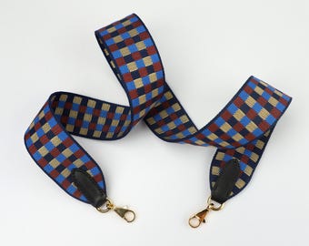 May include: A blue, red, and yellow checkered fabric strap with gold-toned metal clasps. The strap is designed to be used as a replacement shoulder strap for a handbag.