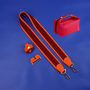 Może przedstawiać: Pasek w jasno pomarańczowe i bordowe paski z pozłacanymi okuciami i dwoma metalowymi klamrami. Pasek znajduje się na niebieskim tle z małą różową torebką.