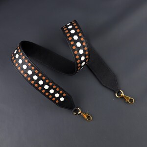 May include: Black bag strap with a geometric pattern of white and brown hexagons. The strap has gold-colored metal clasps on each end. The strap is made of a durable material and is designed to be attached to a bag.
