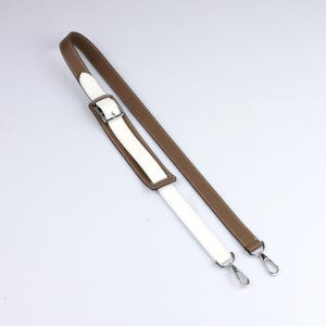May include: A brown and white leather bag strap with silver-tone hardware. The strap features a white central section with brown trim and a matching brown strap. The strap has a buckle and two metal clasps.