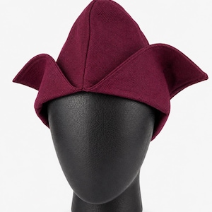 May include: A burgundy-colored hat with a unique, folded design. The hat features a pointed crown and wide, wing-like flaps extending from the sides. The hat is displayed on a black mannequin head against a white background.