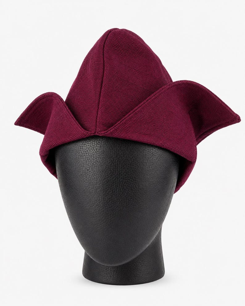 May include: A burgundy-colored hat with a unique, folded design. The hat features a pointed crown and wide, wing-like flaps extending from the sides. The hat is displayed on a black mannequin head against a white background.