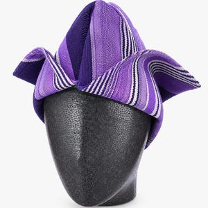 May include: A purple and white striped head wrap, displayed on a black mannequin head. The head wrap features a folded design with varying shades of purple and white stripes. The fabric appears to be woven.