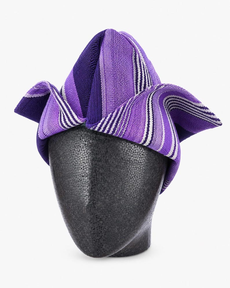 May include: A purple and white striped head wrap, displayed on a black mannequin head. The head wrap features a folded design with varying shades of purple and white stripes. The fabric appears to be woven.