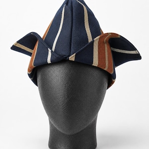 May include: A navy blue, brown, and white striped hat with a unique folded design. The hat is displayed on a black mannequin head, showcasing its shape and style. The hat's design features vertical stripes.