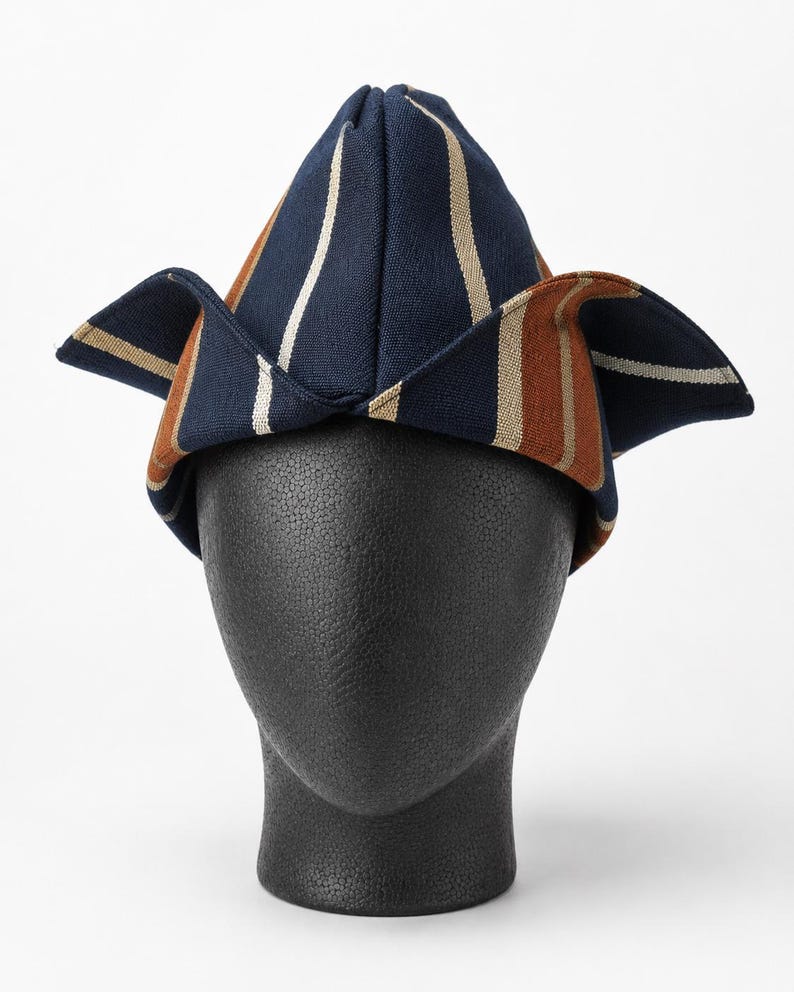 May include: A navy blue, brown, and white striped hat with a unique folded design. The hat is displayed on a black mannequin head, showcasing its shape and style. The hat's design features vertical stripes.
