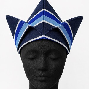 May include: A navy blue, peaked hat with a geometric design. The hat features alternating stripes of white, light blue, and dark blue. The hat is displayed on a black mannequin head.
