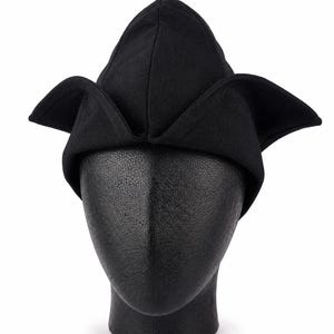 May include: A black hat with a unique, pointed crown and wide, folded brims. The hat is displayed on a black mannequin head against a white background. The hat's design is reminiscent of a medieval or Renaissance style.