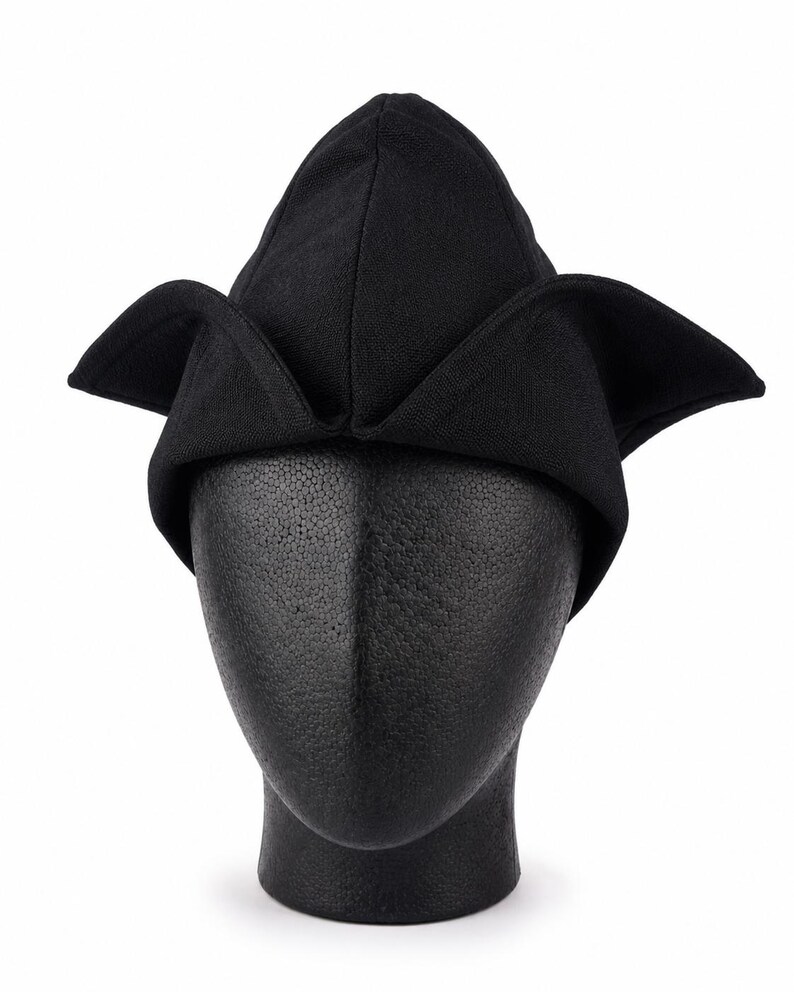 May include: A black hat with a unique, pointed crown and wide, folded brims. The hat is displayed on a black mannequin head against a white background. The hat's design is reminiscent of a medieval or Renaissance style.