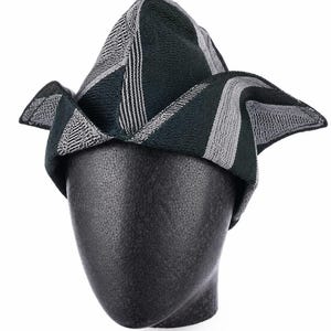 May include: A patterned hat in shades of green, gray, and white, displayed on a black mannequin head. The hat features a geometric design with stripes and angled panels. The brim has a unique, folded shape.