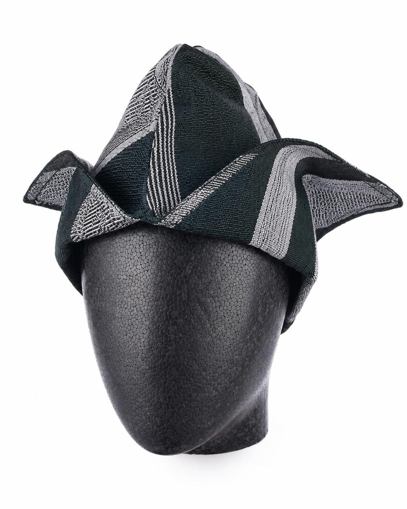 May include: A patterned hat in shades of green, gray, and white, displayed on a black mannequin head. The hat features a geometric design with stripes and angled panels. The brim has a unique, folded shape.
