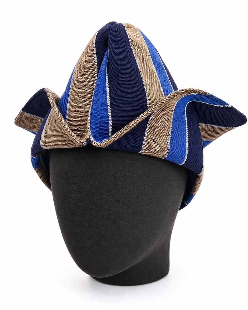 May include: A striped hat in navy blue, light blue, and tan, displayed on a black mannequin head. The hat has a unique, pointed shape with a wide, folded brim. The stripes run vertically, creating a bold pattern.