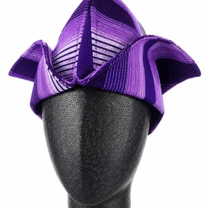 May include: A purple and white striped hat with a unique folded design. The hat is displayed on a black mannequin head. The hat's fabric features varying shades of purple and white stripes, creating a visually striking pattern. The hat's shape is angular and modern.