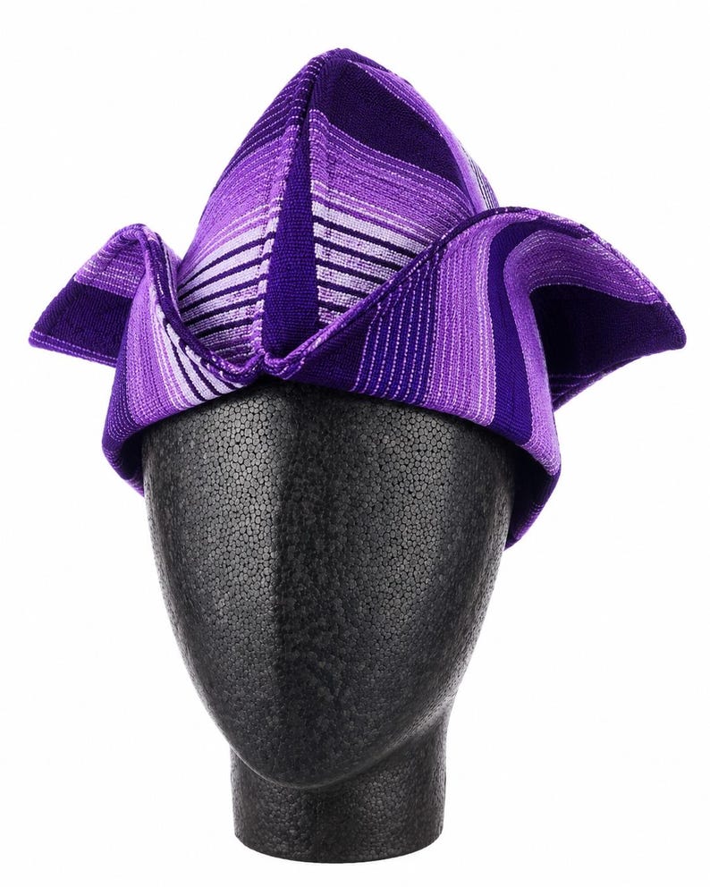 May include: A purple and white striped hat with a unique folded design. The hat is displayed on a black mannequin head. The hat's fabric features varying shades of purple and white stripes, creating a visually striking pattern. The hat's shape is angular and modern.