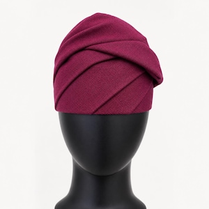 Peut inclure: Un chapeau de style turban bordeaux avec un design pliss&eacute;. Le chapeau, fait de tissu textur&eacute;, est pr&eacute;sent&eacute; sur une t&ecirc;te de mannequin noire. Le design pr&eacute;sente des plis horizontaux qui enveloppent la t&ecirc;te.