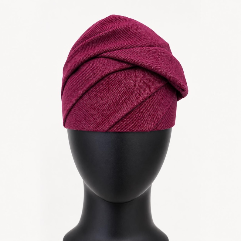Peut inclure: Un chapeau de style turban bordeaux avec un design pliss&eacute;. Le chapeau, fait de tissu textur&eacute;, est pr&eacute;sent&eacute; sur une t&ecirc;te de mannequin noire. Le design pr&eacute;sente des plis horizontaux qui enveloppent la t&ecirc;te.