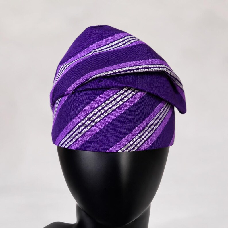 Peut inclure: Un turban &agrave; rayures violettes et blanches. Le tissu est pli&eacute; et drap&eacute; pour cr&eacute;er un couvre-chef &eacute;l&eacute;gant. Les rayures sont un m&eacute;lange de violet et de blanc, cr&eacute;ant un motif visuellement attrayant. Le turban est pr&eacute;sent&eacute; sur une t&ecirc;te de mannequin noire.