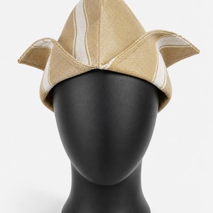 May include: A tan and white striped hat with a unique, angular design. The hat is displayed on a black mannequin head, showcasing its shape and texture. The hat's fabric appears to be woven, with a structured form.