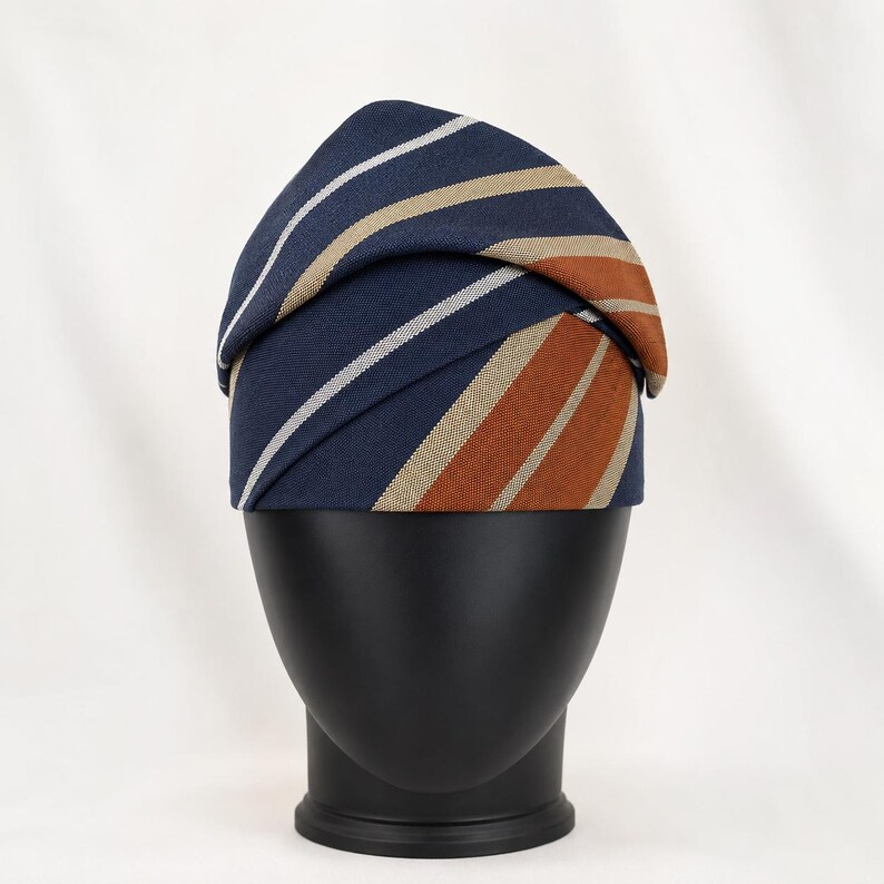 Peut inclure: Un foulard de t&ecirc;te bleu marine avec des rayures blanches, beiges et orange. Le tissu est pli&eacute; et drap&eacute; sur une t&ecirc;te de mannequin noire. Le foulard est un accessoire &eacute;l&eacute;gant.