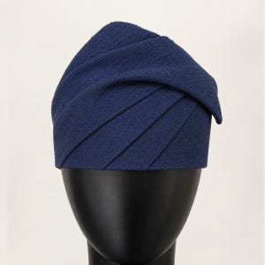 Peut inclure: Un chapeau bleu marine avec un design pliss&eacute; unique. Le tissu du chapeau est de couleur unie, et les plis cr&eacute;ent un effet textur&eacute; et superpos&eacute;. Le chapeau est pr&eacute;sent&eacute; sur une t&ecirc;te de mannequin noire, mettant en valeur sa forme et son style.