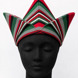 May include: A colorful, striped hat with a unique, angular design. The hat features red, green, black, white, and gray stripes. The hat is displayed on a black mannequin head against a white background.