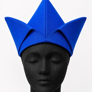 May include: A vibrant blue hat with a distinctive, angular design. The hat features a central, pointed crown and four outward-facing, triangular flaps. The hat is made of a smooth, solid-colored fabric and is displayed on a black mannequin head.